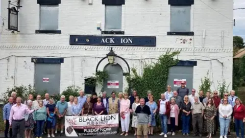A boarded-up white building with faded signage for “Black Lion” stands in the background, with ivy growing along one side. In front, a large group of people gathers closely together, holding a banner that reads “Save Black Lion Pub & Hub – Community Owned.” The scene suggests a community campaign or protest to support and reopen the pub.