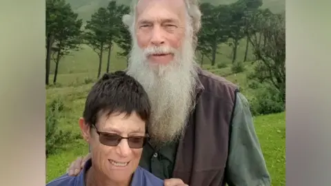 Rodney Saunders stands behind Rachel. Rodney has a long grey beard while Rachel has dark hair and is wearing sunglasses