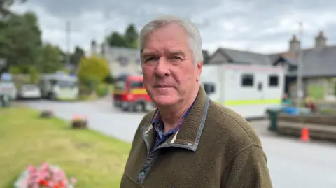 Sandy McCook is standing outside. He has grey hair and is wearing a green fleece over a tartan shirt.