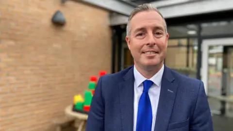 The Minister Paul Givan pictured in 2024. He has a bright blue tie, navy suit and white shirt. He has shirt grey hair and smiles at the camera.