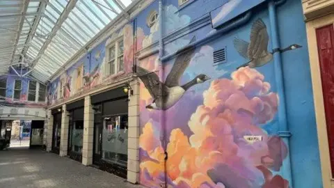 The mural painted on the wall inside Lowther Arcade. A large section of wall shows two geese flying through a blue sky with their wings outstretched. behind them are clouds which are coloured pink, orange and purple as if catching the light from the sun. The mural stretches out above shop fronts inside the arcade.