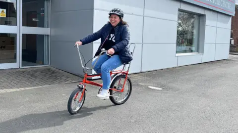 A woman with long dark hair, wearing a cycling helmet, laughs as she rides a red chopper bicycle in front of a building.