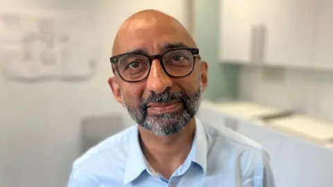 Dr Saket Priyadarshi - he has glasses, a beard and a light blue shirt on. He is standing in a medical room and looking directly into the camera.