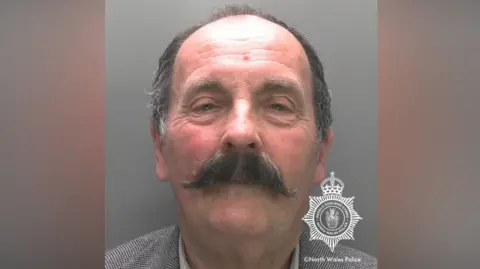 North Wales Police A man with dark and grey hair and a mainly dark moustache is looking at the camera. It includes a North Wales Police watermark.
