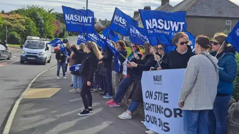 Union members on a picket line waving blue flags with the NASUWT logo beside a road. 