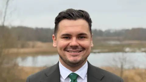 A man with slicked back hair and wearing a grey blazer, white shirt and green tie smiles in front of a background featuring an out-of-focus lake and greenery
