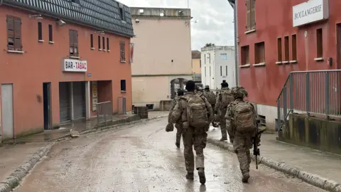An army exercise is carried out with a group of soldiers running down a muddy road - with a boulangerie sign on the right and a tabac sign on the left. They are carrying backpacks.