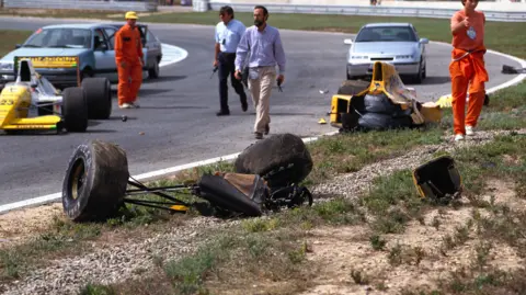 Getty Images A yellow racing car is in the half following a crash Pieces of the car are scatted across part of the grey track and on the grass to the right. People are standing and walking around the area.