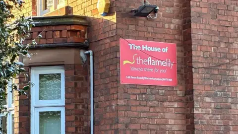 A sign on the property where the agency is based reads "the House of the Flame Lily, always there for you". The brick building has sunlight on it and there are blinds at the windows.