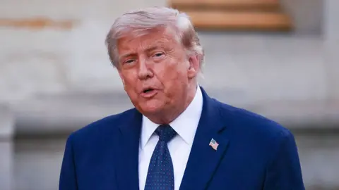 Trump is seen speaking. He is wearing a blue suit and a tie with the American flag as a pin on it 