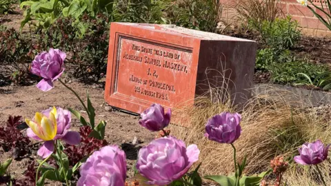 A block taken from a building. It is inscribed with details about who the brick was laid by. In the foreground are purple flowers, with grasses and other plants surrounding the block.