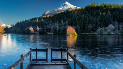 Andrew Briggs A bridge sits on a frozen loch. Wintry looking trees  are visible in the distance and at the top is a snow covered mountain peak