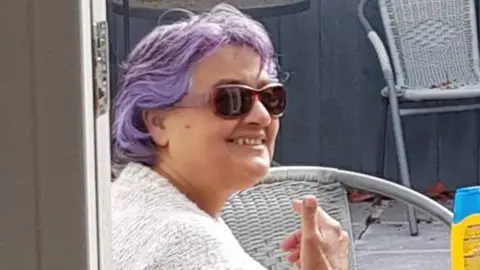 Family handout Bernadette smiling at the camera. She is wearing sun glasses and is sat in a garden. She has purple hair. 