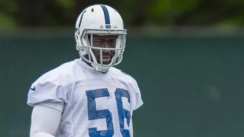 He is wearing his American football uniform, but the image is cut just below his shoulders. He is wearing a white helmet with a blue stripe down the middle and a white tshirt with blue detailing