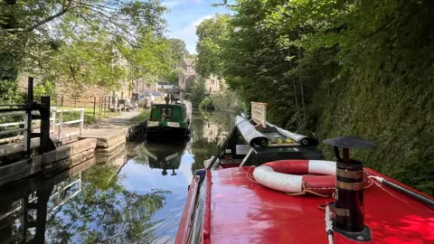 Olivia Richwald/BBC The picture has been taken onboard a red narrow boat cruising along the canal. You can see a red roof with a chimney and red and white life ring. A green narrow boat is in the distance.