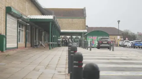 A brick supermarket with green arches is visible on the left behind black boulders, to the right are parking bays and some cars lined up