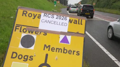A roadside sign points towards the Royal Cornwall Show, with a paper notice taped over it reading “RCS 2026 CANCELLED.” In the background, a line of cars queues along a wet road.