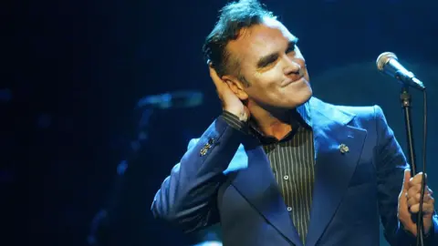 Morrissey, wearing a blue suit jacket, holds one hand to his head as he performs on stage