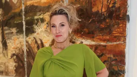A woman wearing a light green dress. She has her hair up and is wearing gold earrings and a necklace.