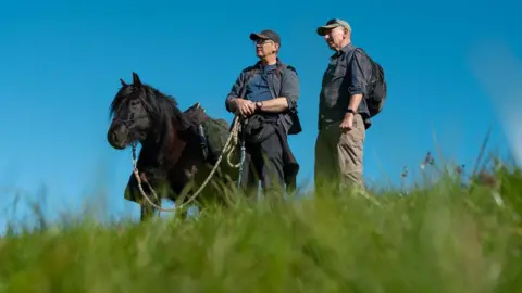sesmlphotography Roger Sewill standing on Hod Hill with his pony Scarlet and another man. They are looking into the distance, away from the camera. It is a clear day.