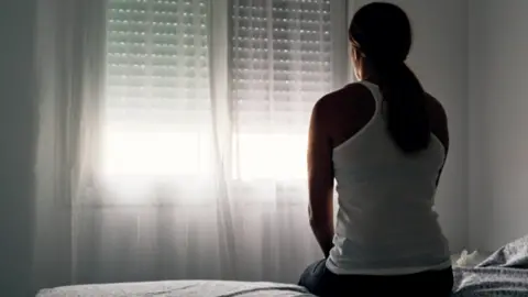 Alvaro Medina Jurado A woman sits on a bed with her back to the camera, looking towards a window