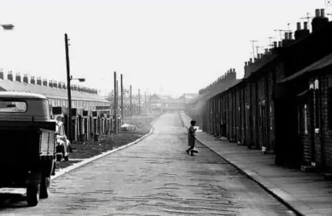 Eileen Doyle Dawson  A black and white image of rows and rows of terraced miners' houses with a van and a single car parked up and a woman carrying a brush walking across the road.