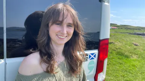 Danielle Stewart has long, brown hair and is wearing a green top. She is standing outside next to the rear of her campervan on a sunny day. There is a large grassy area in the background.