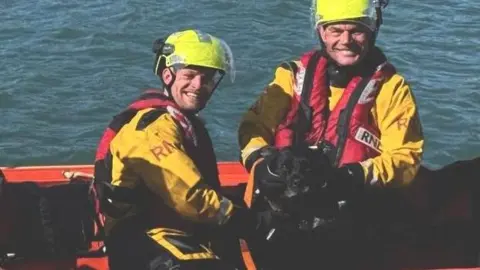 Two RNLI liveboat crew smile toward the camera wearing yellow uniforms with red life jackets in a boat and holding on to a black dog.