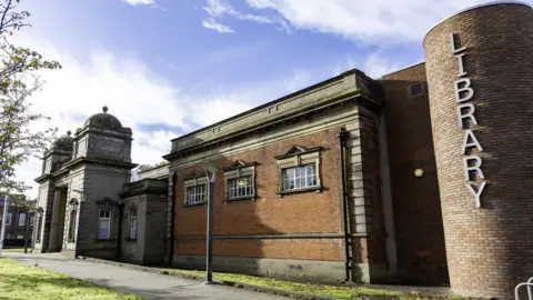 Gateshead Library An external shot of Gateshead Central Library. Most of the building is made of red brick. The large entrance is built into an archway topped with two domes.