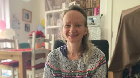 Amy Morris is pictured sitting on a chair in a living area in her home. She is smiling and is wearing a grey knitted jumper with pink, green and blue lines across it. Behind her is an out-of-focus table and chairs. There are pictures on the walls.