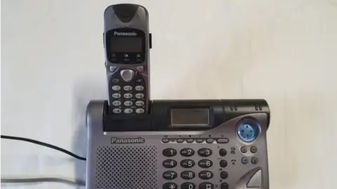 A grey phone from the 1970s or 80s, which has a receiver, number buttons and a screen for an answerphone. 