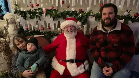 Family handout Three-year-old Ralf admires the Christmas decorations as he meets Santa for the first time without shielding. Father Christmas in sat in the middle, with one arm around Daisy, who is cuddling Ralf, who is looking up and dressed in a black beanie and green winter coat and gloves, and another arm around Ralf's dad Doug, dressed in a red and black checked shirt and jeans. Strings of baubles and fake icicles are behind them