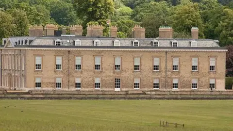 Getty Images A long, rectangular old stone building with multiple windows and several chimney stacks on the roof, set behind a wide green lawn with trees and woodland in the background.
