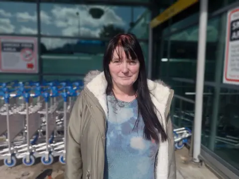 A woman with black hair wearing a blue blouse and coat standing outside an airport in front of luggage trolleys