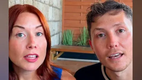 BBC Bec and Sam sit side by side indoors speaking into black microphones. A concrete block wall, wooden panelled screen and potted plants are visible behind them.