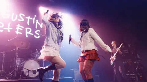 AJ Stark Two girls dancing on a stage wearing sunglasses. One is wearing a blue shirt and a black tie with a white frilly skirt and black boots. The other is wearing a white shire with a red frilly skirt, red lace tights and black boots. They are both holding microphones. There is a set of drums to the back of the stage and some writing that is cut off. There is a man to the right holding a guitar. 