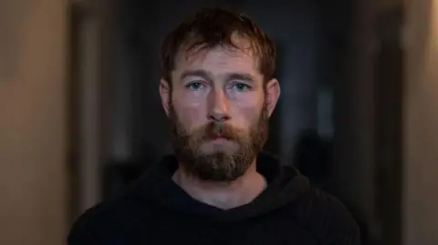 Chris Garrett has brown short hair and a beard. He's wearing a black hoodie.