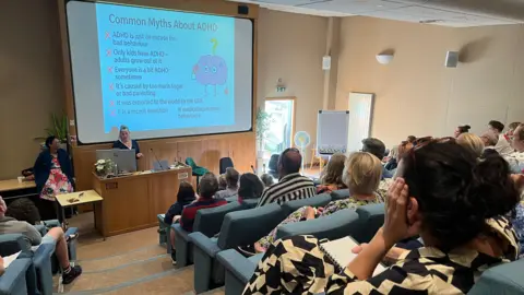 A lecture theatre with about 100 people in it facing the front. On a screen behind a desk is a presentation titled "Common Myths About ADHD", featuring a cartoon brain. Rachel Boss is presenting a lecture at the podium.
