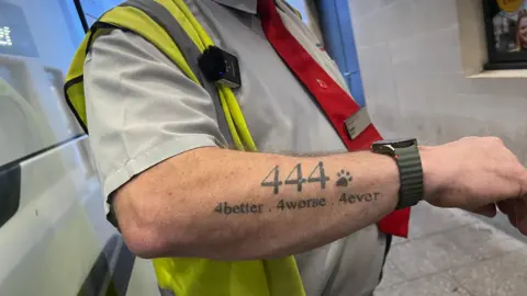 A man is showing a tattoo on his forearm which says "444. 4better. 4worse. 4ever". He is wearing a smart watch too, and a short sleeve shirt and high visibility jacket.