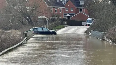 A black mini part-submerged in floodwater on a road over a bridge