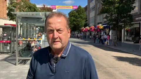 A middle aged man in a blue polo shirt stands in a busy shopping street in the centre of Exeter.