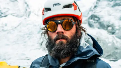 Blue Door Productions/Stan Gaskell Mitch Hutchcraft appears with a long thick dark beard, wearing protective sunglasses and a waterproof jacket, standing in front of snow or ice.