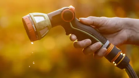 A hand holding the head of a hosepipe sprayer. The head of the hosepipe is black and orange and there are water droplets dripping from it.