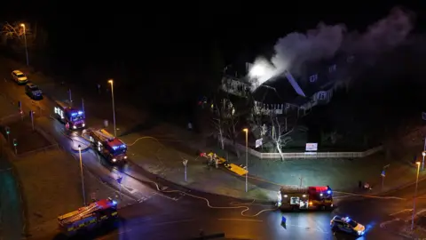 An aerial shot shows four fire engines parked at the side of the road and a large pub building is seen with smoke coming out its roof. It is dark and the vehicles have their lights on and there are fire hoses across the road and pavement