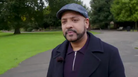 A man in a purple collared shirt, a navy blue coat, a blue flat cap. He has a beard and moustache. He stands in a park, with trees and grass in the background.