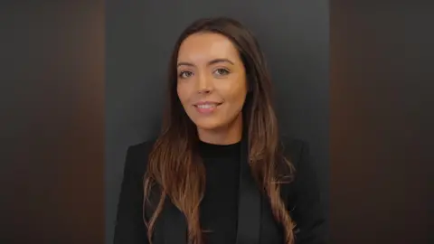 Phoenix Law Victoria smiling at the camera. She has long brown hair and brown eyes. She is wearing a black top under a black blazer and is standing in front of a black wall.