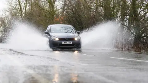PA Media A dark grey sedan car with full beams on sprays standing water either side of it as it goes down a wet, tree-lined road.