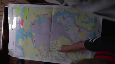 Ella Hibbert's finger pointing at a location on a world map which shows the arctic circle and multiple green, yellow, orange and pink post-it notes attached
