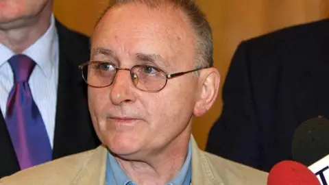 A man in a blue shirt and beige jacket looks to the left of the camera. Two other people dressed in suits are standing behind him, although their faces are not visible. The man has short dark hair, which is turning grey, and is wearing glasses. Several microphones are also visible to his right, pointed in his direction. 