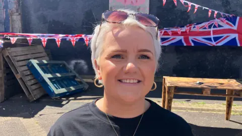  Stacey Graham has short, cropped blond hair and wears sunglasses on her head and a black t-shirt. She is standing outside and there is Union Jack bunting and flags in the background. 